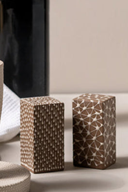 A styled tabletop scene with a dark wine bottle, a textured beige drinking cup and matching coaster, two patterned salt and pepper shakers, a cork, and a loaf of bread wrapped in a white cloth against a plain warm beige background.