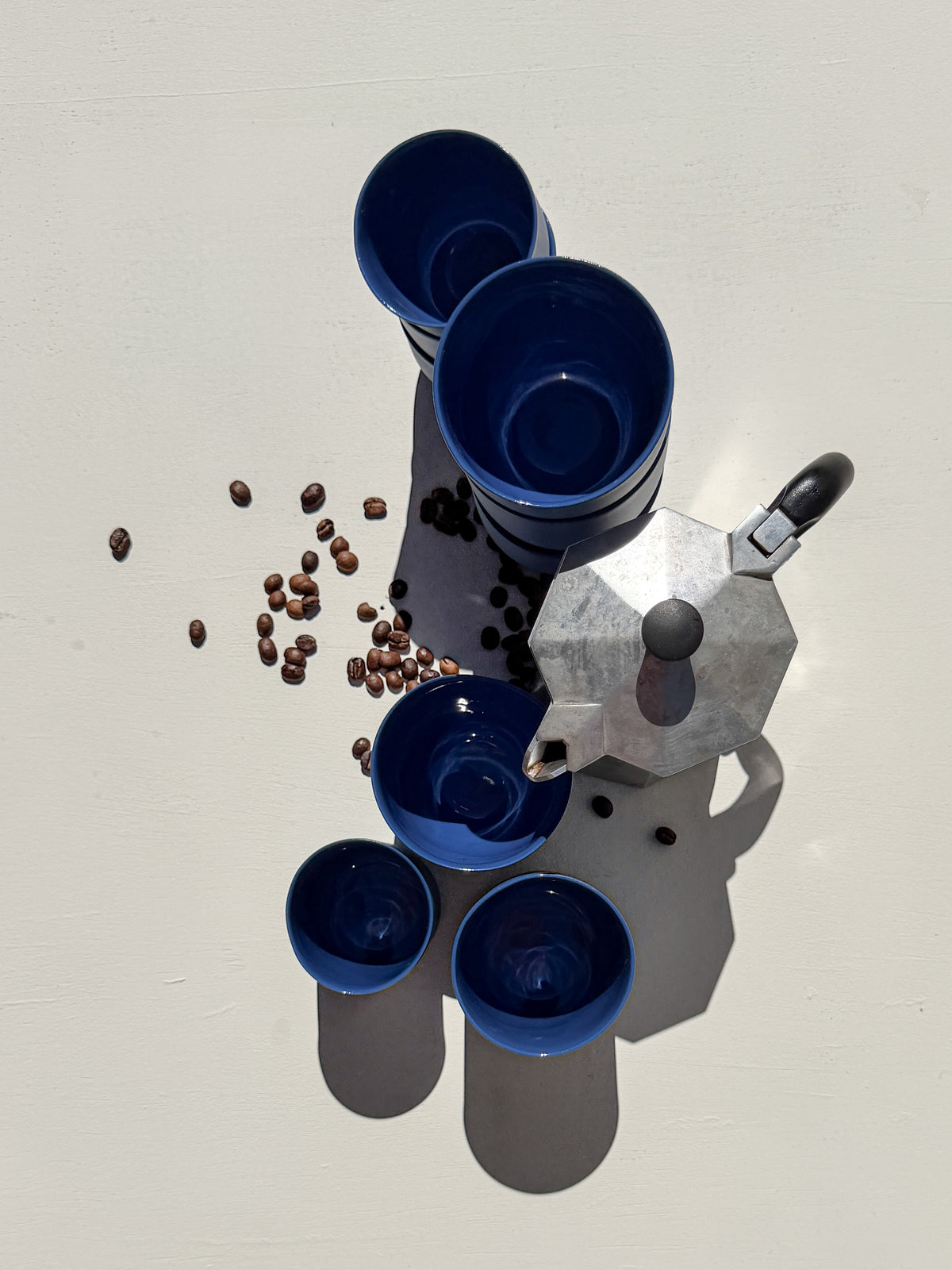 כוסות קרמיקה בגוון כחול מרוקו במבט מלמעלה, לצד מקנקן קפה מתכתי ופולי קפה, על משטח בהיר | Ceramic cups in Morocco blue seen from above, beside a metal coffee maker and coffee beans, on a light surface