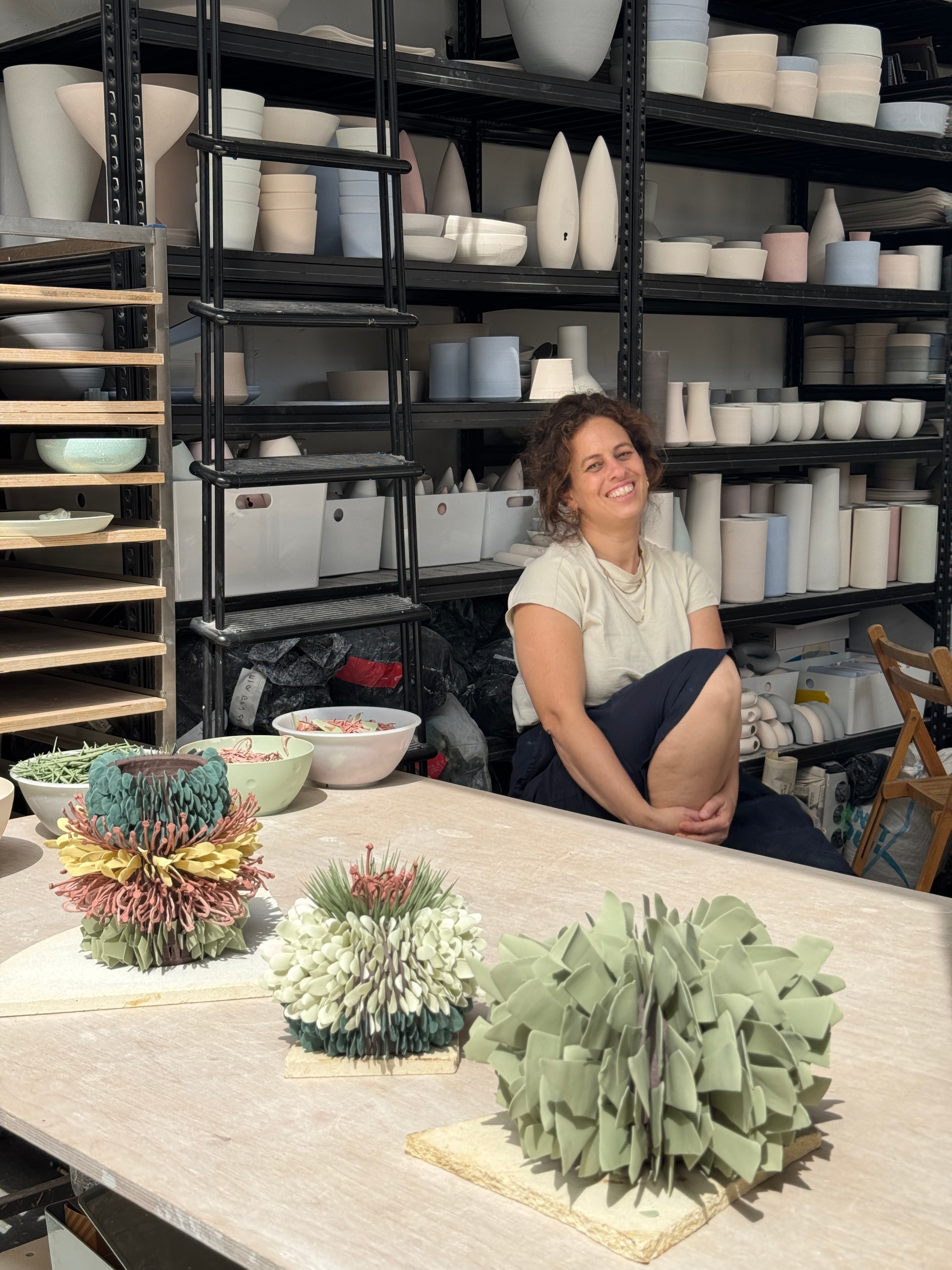 A smiling woman sits on a table in a pottery studio, surrounded by shelves of ceramic vessels. In the foreground on the table are three sculptural, textured succulent-like plant arrangements in muted green, white, and multicolor tones.
